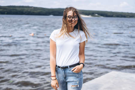 Portrait of a beautiful young woman in stylish glasses. Girl in white tshirt posing on the background of the lake landscape.の写真素材