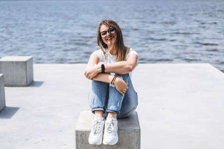 Young woman with long hair in stylish glasses posing on the concrete shore near the lake. Girl dressed in jeans and t-shirt smiling and looking away.の写真素材