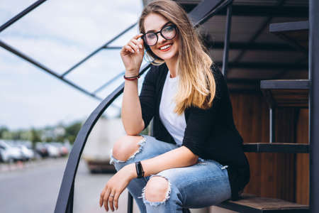 Beautiful girl with long hair and glasses sitting on metal stairs on the wooden background of house with vertical boards. Woman smiling and looking at camera.の写真素材