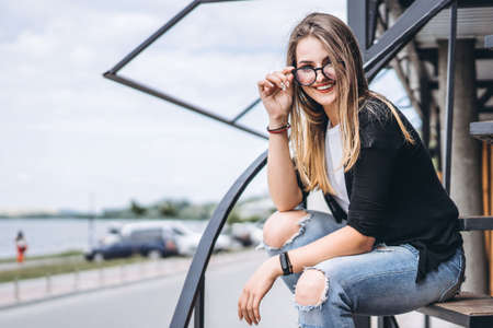 Beautiful girl with long hair and glasses sitting on metal stairs on the wooden background of house with vertical boards. Woman smiling and looking at camera.の写真素材