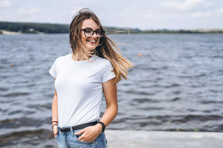 Portrait of a beautiful young woman in stylish glasses. Girl in white tshirt posing on the background of the lake landscape.の写真素材