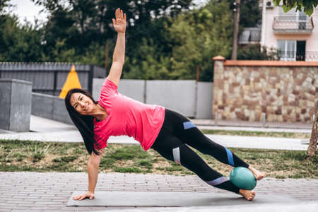 Sporty young woman with long hair doing stretching exercises with a special ball on the street, on the background of trees. Active lifestyle, yoga concept.の写真素材