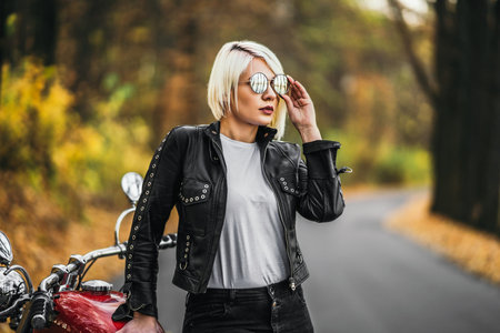 Pretty blonde biker girl in sunglasses with red motorcycle on the road in the forest with colorful blurred background.の写真素材