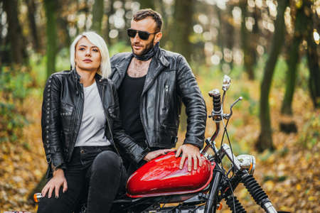 Pretty couple near red motorcycle on the road in the forest with colorful blured background. Relationship concept.の写真素材