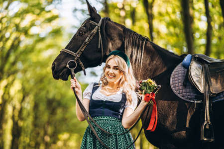 Pretty blonde in traditional dress  walking with big black horse in the forest. Animal and people frienship concept.の写真素材