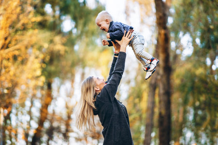 Happy mother with her little baby son have fun in the city park with colorful leaves around in...