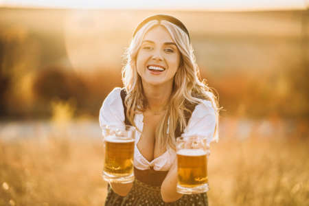 Pretty happy blonde in dirndl, traditional festival dress, holding two mugs of beer outdoors in the field with blurred background. Oktoberfest, St. Patrickâs day, international beer day concept.の写真素材