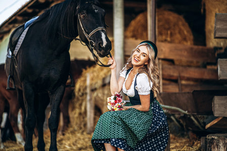 Pretty blonde in traditional dress  take care of big black horse at the farm. Animal and people frienship concept.の写真素材