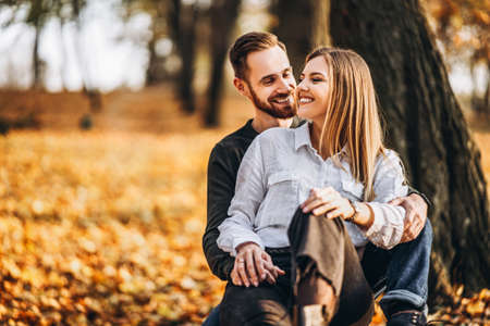 A young loving couple sitting on a wooden bench in the forest. Man and woman hugging and smiling on the background of autumn trees.の写真素材
