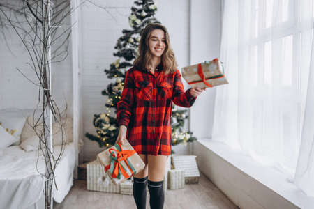 Christmas, New Year. Pretty woman in shirt and socks walking at home with christmas gift box, new year tree behindの写真素材