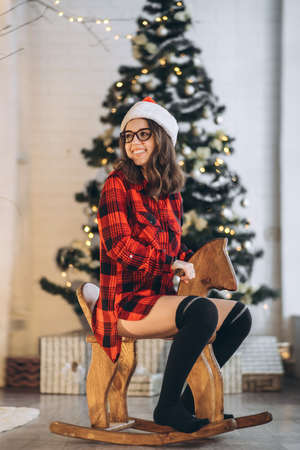 Christmas, New Year. Pretty woman in shirt and socks have fun riding on the wooden swing horse toy, new year tree behindの写真素材