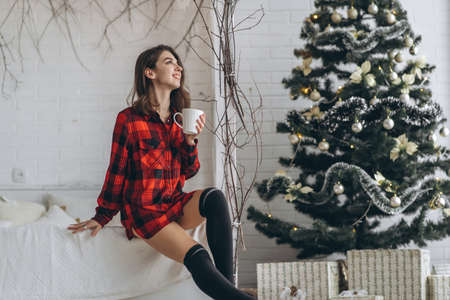 Pretty brunette girl in bed wearing red shirt and warm socks with cup of coffee and christmas lights behindの写真素材