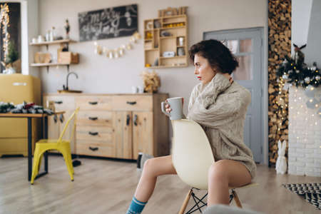 Pretty and happy brunette sitting in warm sweater on the chair, holding cup of hot beverage with christmas lights behindの写真素材