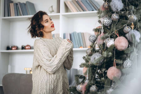 Christmas, New Year. Pretty and happy brunette in warm sweater decorating christmas tree at homeの写真素材