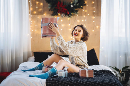 Happy and pretty brunette on the bed with gift boxes around her. New Year, Christmas, birthday,...