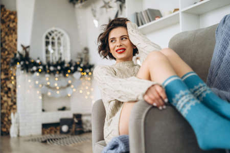 Pretty and happy brunette sitting in warm sweater on the chair with christmas lights behindの写真素材