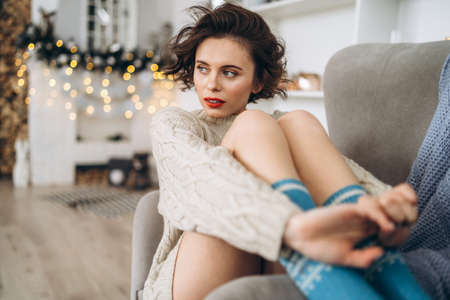 Pretty and happy brunette sitting in warm sweater on the chair with christmas lights behindの写真素材