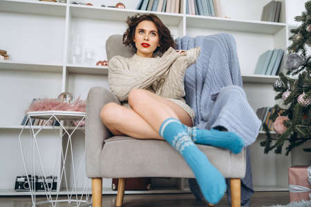 Pretty and happy brunette sitting in warm sweater on the chair with christmas lights behindの写真素材