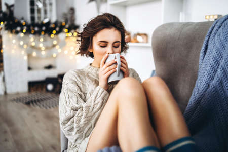 Pretty and happy brunette sitting in warm sweater on the chair, holding cup of hot beverage with christmas lights behindの写真素材