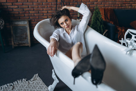 Portrait of a beautiful brunette lying in an empty bathroom holding legs up, wearing a white shirt...
