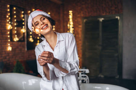 A young brunette sitting on the edge of the bath in Ð¡hristmas hat holding burning sparkler firework bengal light in handsの写真素材