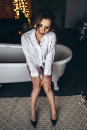 A young brunette sitting on the edge of the bath and looks at the camera. Woman wearing a white...