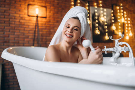 A beautiful young woman posing in studio. Woman lying in a white bath with a towel on her head. A red brick wall with yellow garlands on the background.の写真素材