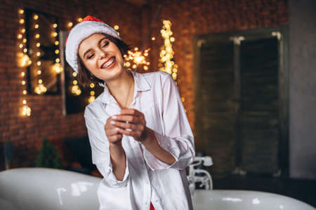 A young brunette sitting on the edge of the bath in Ð¡hristmas hat holding burning sparkler firework bengal light in handsの写真素材