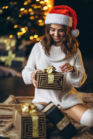 Christmas, New Year. Pretty woman in warm sweater, socks and christmas hat, sitting on the floor at home with gift boxes and new year tree and lights behindの写真素材