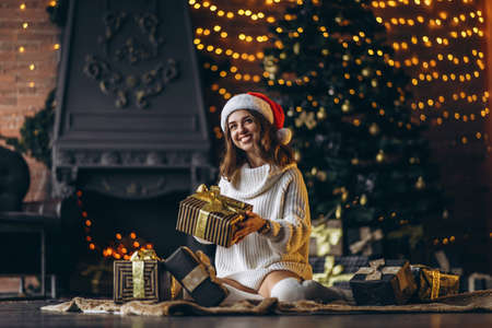 Christmas, New Year. Pretty woman in warm sweater, socks and christmas hat, sitting on the floor at home with gift boxes and new year tree and lights behindの写真素材