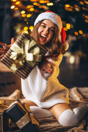 Christmas, New Year. Pretty woman in warm sweater, socks and christmas hat, sitting on the floor at home with gift boxes and new year tree and lights behindの写真素材