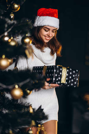 Christmas, New Year. Pretty woman in warm sweater, socks and christmas hat, decorating new year tree and holding gif boxの写真素材