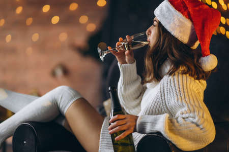 Christmas, New Year. Pretty woman in warm sweater, socks and christmas hat, sitting on the chair with champagne and new year tree and lights behindの写真素材