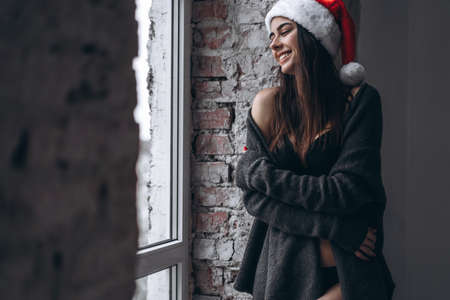 Christmas, New Year. Pretty girl in lingerie, sweater and Christmas hat posing indoors near windowの写真素材