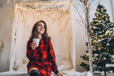Pretty brunette girl in bed wearing red shirt and warm socks with cup of coffee and christmas lights behindの写真素材