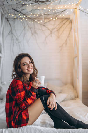 Pretty brunette girl in bed wearing red shirt and warm socks with cup of coffee and christmas lights behindの写真素材