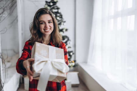 Christmas, New Year. Pretty woman in shirt and socks walking at home with christmas gift box, new year tree behindの写真素材