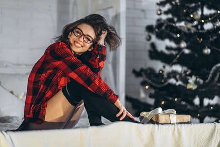 Christmas, New Year. Pretty woman in shirt and socks lying in bed with christmas gift box, new year tree behindの写真素材