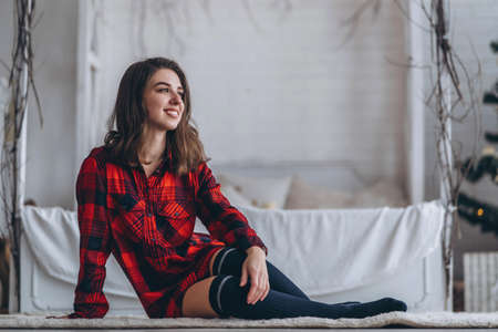Pretty brunette girl sitting on the floor in red shirt and warm socks with christmas tree behindの写真素材