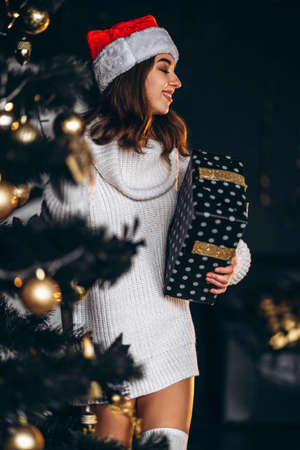 Christmas, New Year. Pretty woman in warm sweater, socks and christmas hat, decorating new year tree and holding gif boxの写真素材