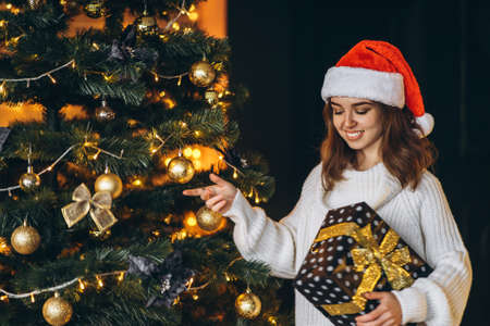 Christmas, New Year. Pretty woman in warm sweater, socks and christmas hat, decorating new year tree and holding gif boxの写真素材