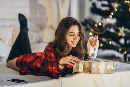 Christmas, New Year. Pretty woman in shirt and socks lying in bed with christmas gift box, new year tree behindの写真素材