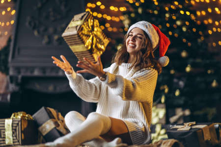 Christmas, New Year. Pretty woman in warm sweater, socks and Christmas hat, sitting on the floor at home with gift boxes and new year tree and lights behindの写真素材