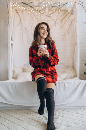 Pretty brunette girl in bed wearing red shirt and warm socks with cup of coffee and Christmas lights behindの写真素材