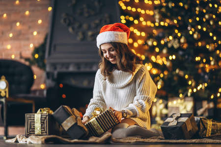 Christmas, New Year. Pretty woman in warm sweater, socks and Christmas hat, sitting on the floor at home with gift boxes and new year tree and lights behindの写真素材