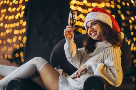 Christmas, New Year. Pretty woman in warm sweater, socks and Christmas hat, sitting on the chair with champagne and new year tree and lights behindの写真素材