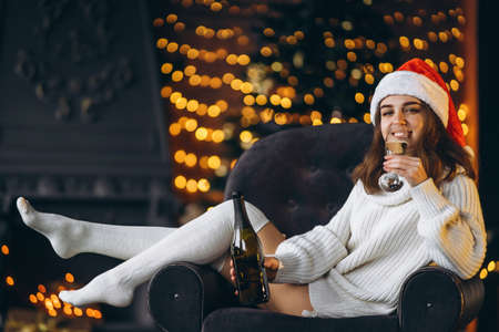Christmas, New Year. Pretty woman in warm sweater, socks and Christmas hat, sitting on the chair with champagne and new year tree and lights behindの写真素材