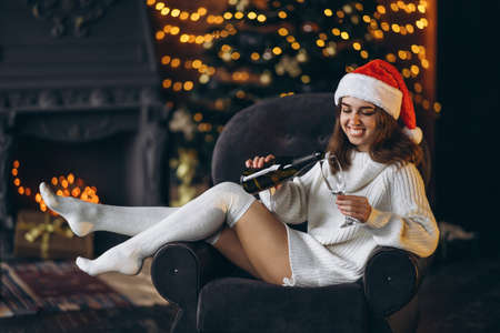 Christmas, New Year. Pretty woman in warm sweater, socks and Christmas hat, sitting on the chair with champagne and new year tree and lights behindの写真素材