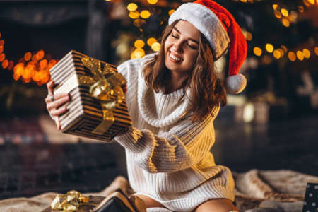 Christmas, New Year. Pretty woman in warm sweater, socks and Christmas hat, sitting on the floor at home with gift boxes and new year tree and lights behindの写真素材