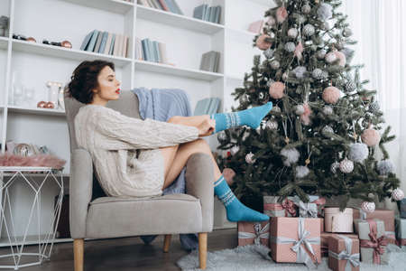 Pretty and happy brunette sitting in warm sweater on the chair with christmas lights behindの写真素材